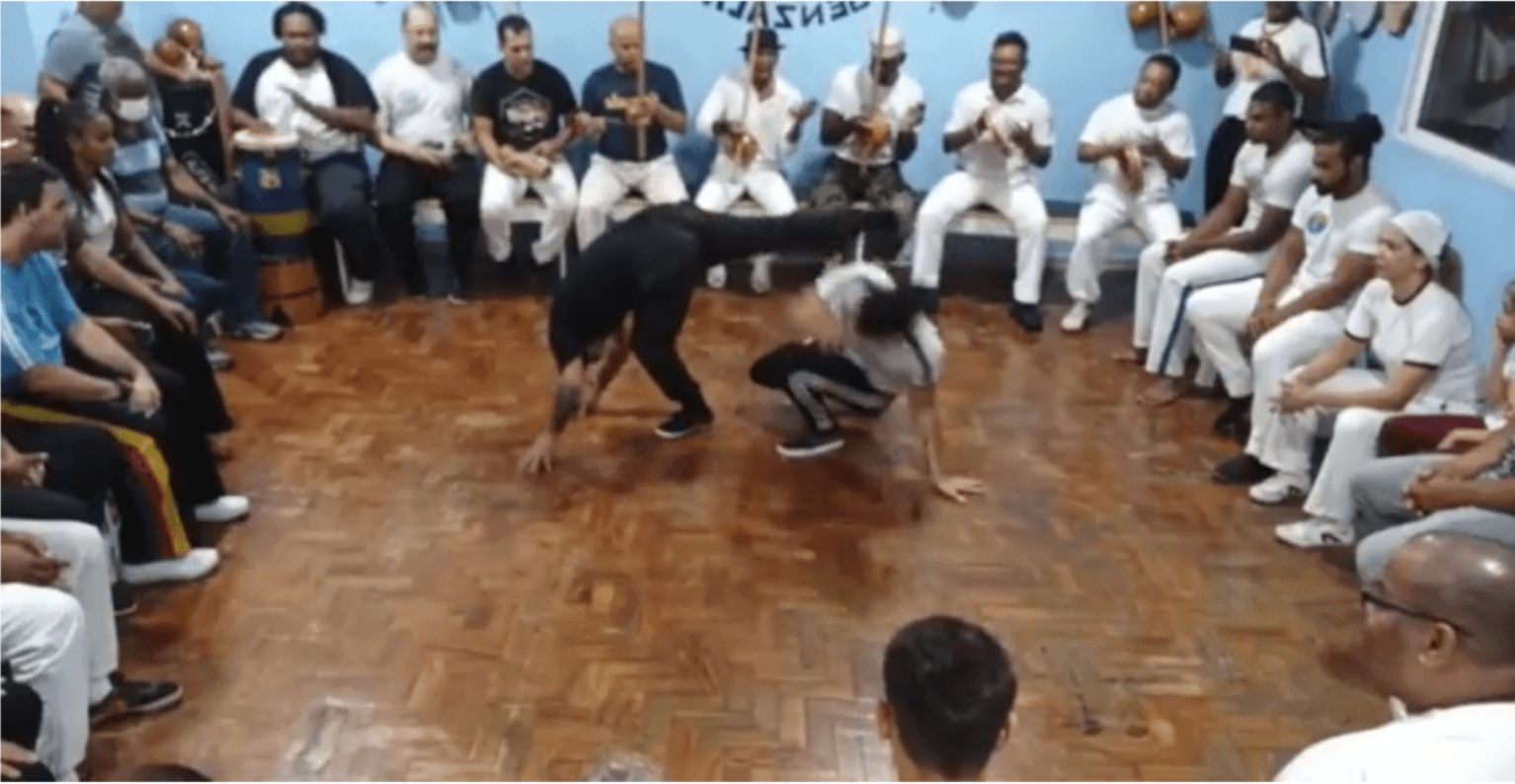 A capoeira circle (roda) in the Senzala de Santos group in Santos, SP, Brazil in December 2021. Author is in white shirt in the centre playing with C.M Val. At the back we see the “bateria,” the eight people playing the instruments led by the berimbau player at the centre. Frame from video by Janaina Bispo Celestino.